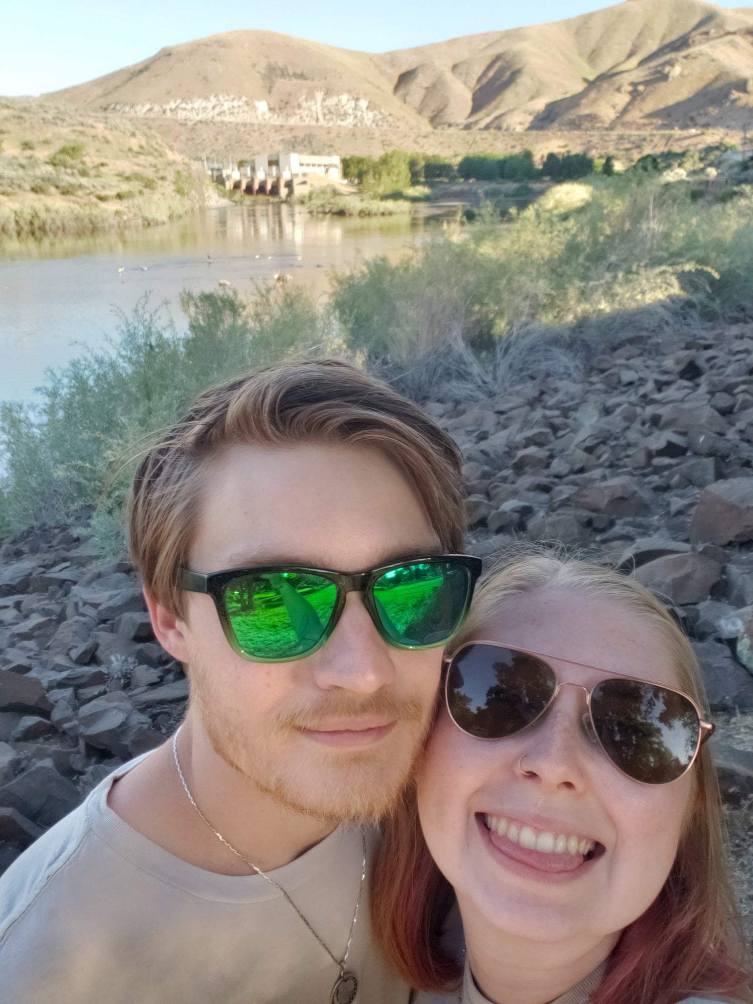 Owners behind Kanna's Botanicals; Aidan & Kailee Poe outside with sunglasses on in front of a black rocky surface with a river and hillside behind them. - Kanna's Botanicals