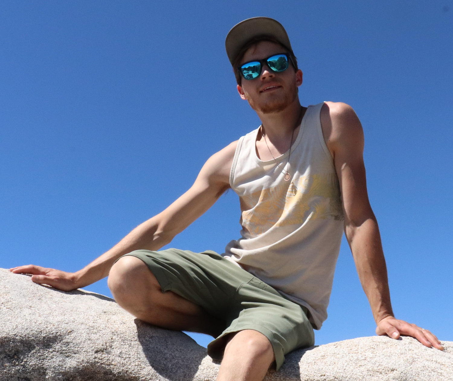 Aidan Poe in a tank top and shorts with a hat and sunglasses on sitting on a large rock surface with a light blue sky. - Kanna's Botanicals