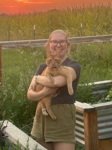 Kailee Poe in a t-shirt and shorts holding an orange cat, Odo, in a garden bed area with a corn field and orange sunset in the background.
 - Kanna's Botanicals